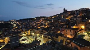 A densely packed hillside town at dusk with illuminated buildings and a prominent church spire. - An elegant retreat overlooking the city of mosaics (Caltagirone)