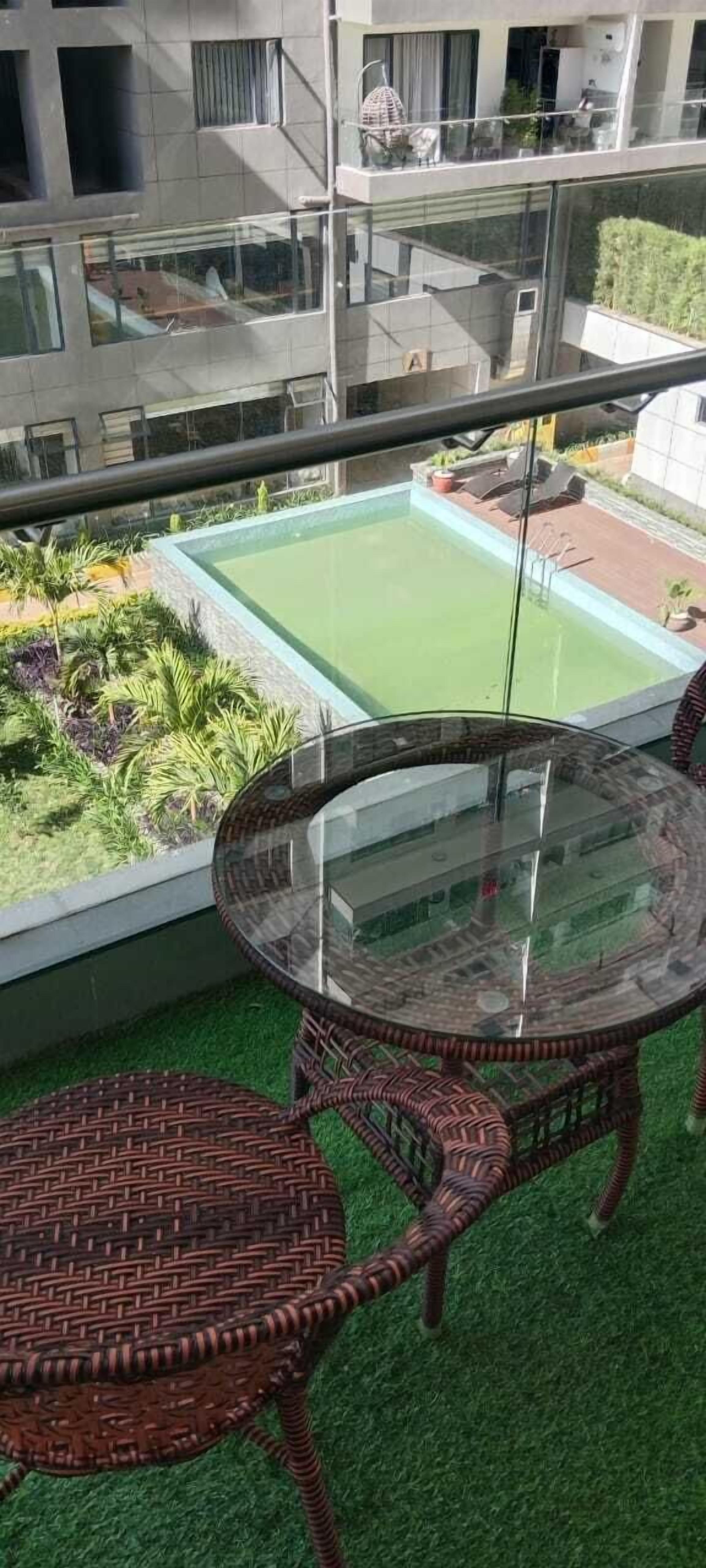A balcony with a glass table and wicker chairs, overlooking a swimming pool and a garden.