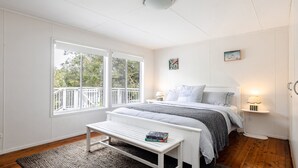 Ein Schlafzimmer mit einem Bett, Nachttischlampen, einer Bank mit Büchern und einem Fenster mit Blick auf Bäume.