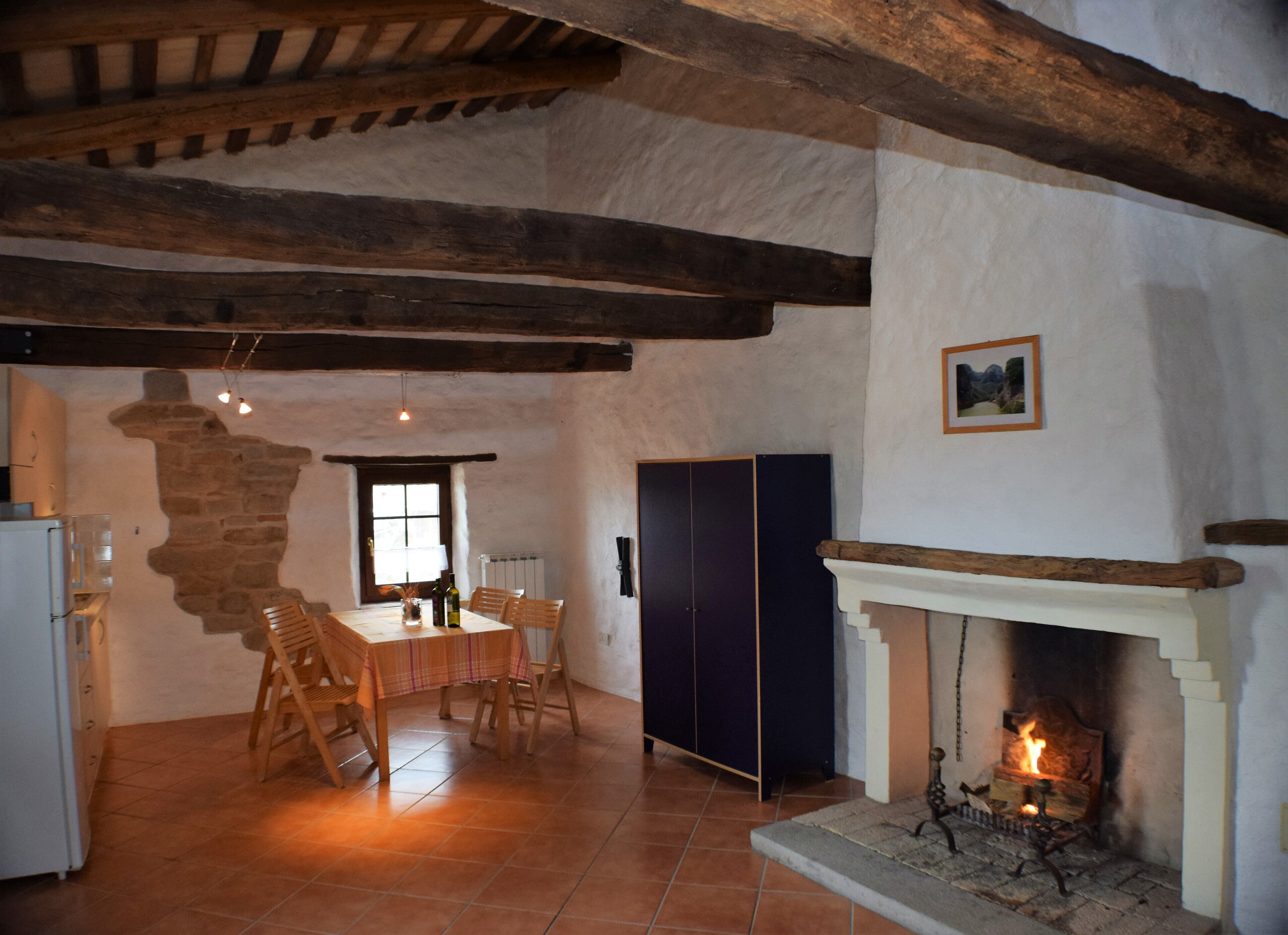 A rustic interior with exposed wooden beams, a fireplace, a dining area with a table and chairs, and a stone wall.
