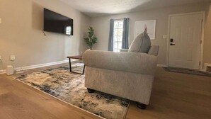 A living room with a sofa, a flat-screen TV mounted on the wall, a window with curtains, and a door. - Stockbridge Home w/ Master Suite on the Main (Stockbridge)