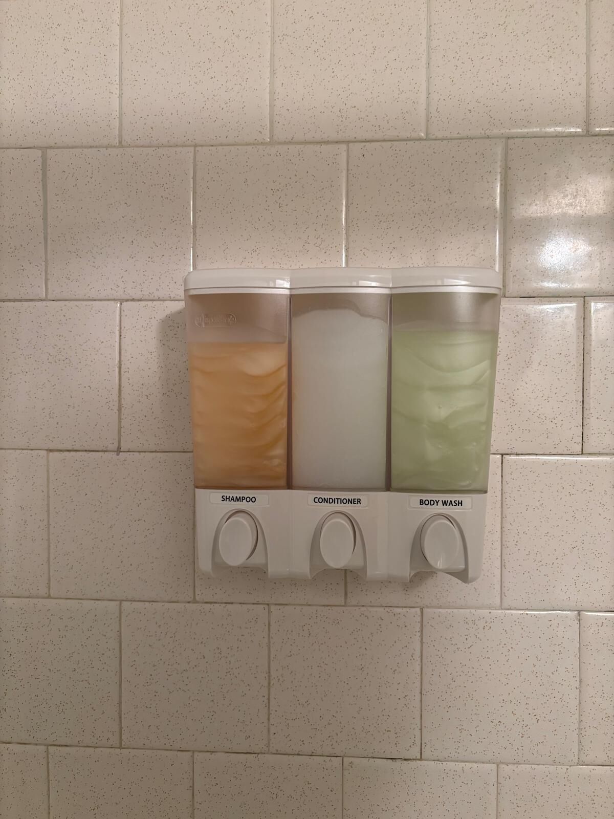 Un distributeur mural doté de trois compartiments étiquetés pour le shampoing, l’après-shampoing et le gel douche.