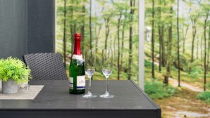 Une bouteille de vin et deux verres sur une table, avec vue sur une forêt par la fenêtre.