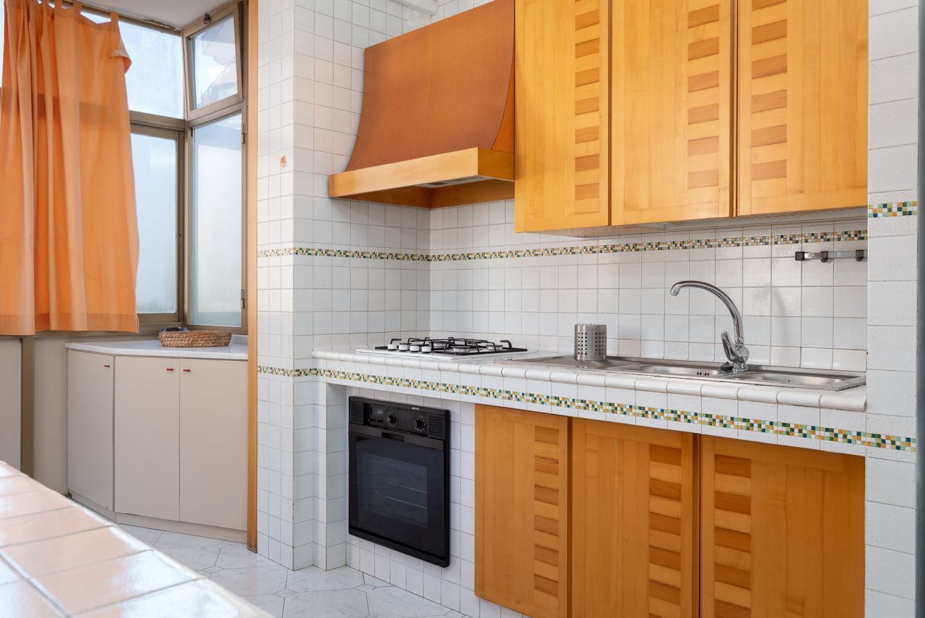 A kitchen with white tiled walls, wooden cabinets, a gas stove, a sink, and a built-in oven.