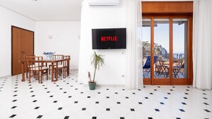 A room with a dining table, chairs, a potted plant, a TV screen displaying 'NETFLIX,' and a view of a balcony with outdoor furniture. - Capo Sereno - Large & Bright apt with terrace (Sorrento)