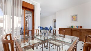 A dining area with a glass-top table, wooden chairs, a vase with blue flowers, and a bookshelf. - Capo Sereno - Large & Bright apt with terrace (Sorrento)