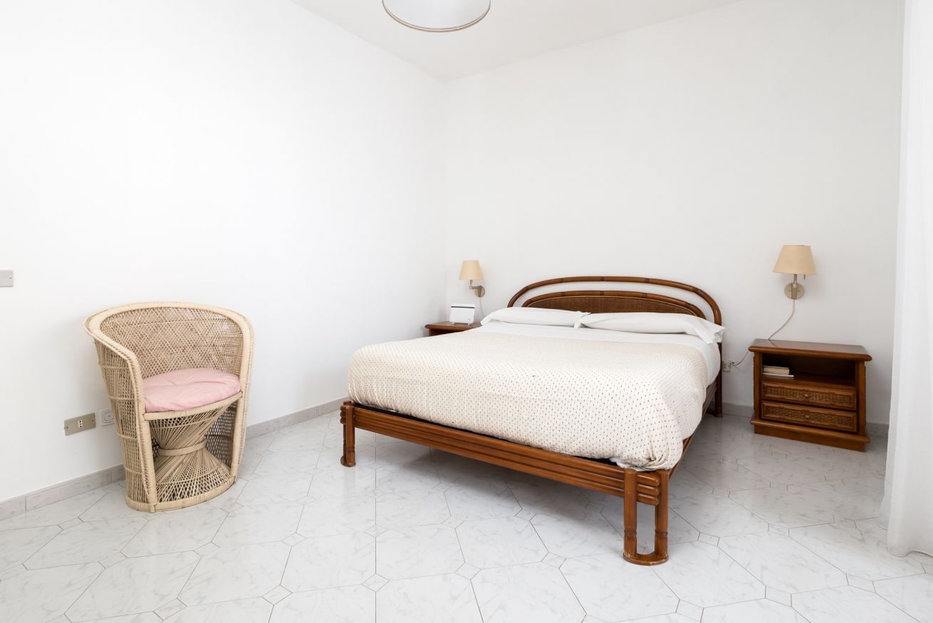 A bedroom with a wooden bed, a wicker chair, a nightstand, and a wall-mounted lamp.