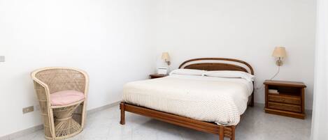 A bedroom with a wooden bed, a wicker chair, a nightstand, and a wall-mounted lamp.