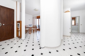 A hotel corridor with a wooden door, white columns, a dining area with a table and chairs, and a tiled floor.