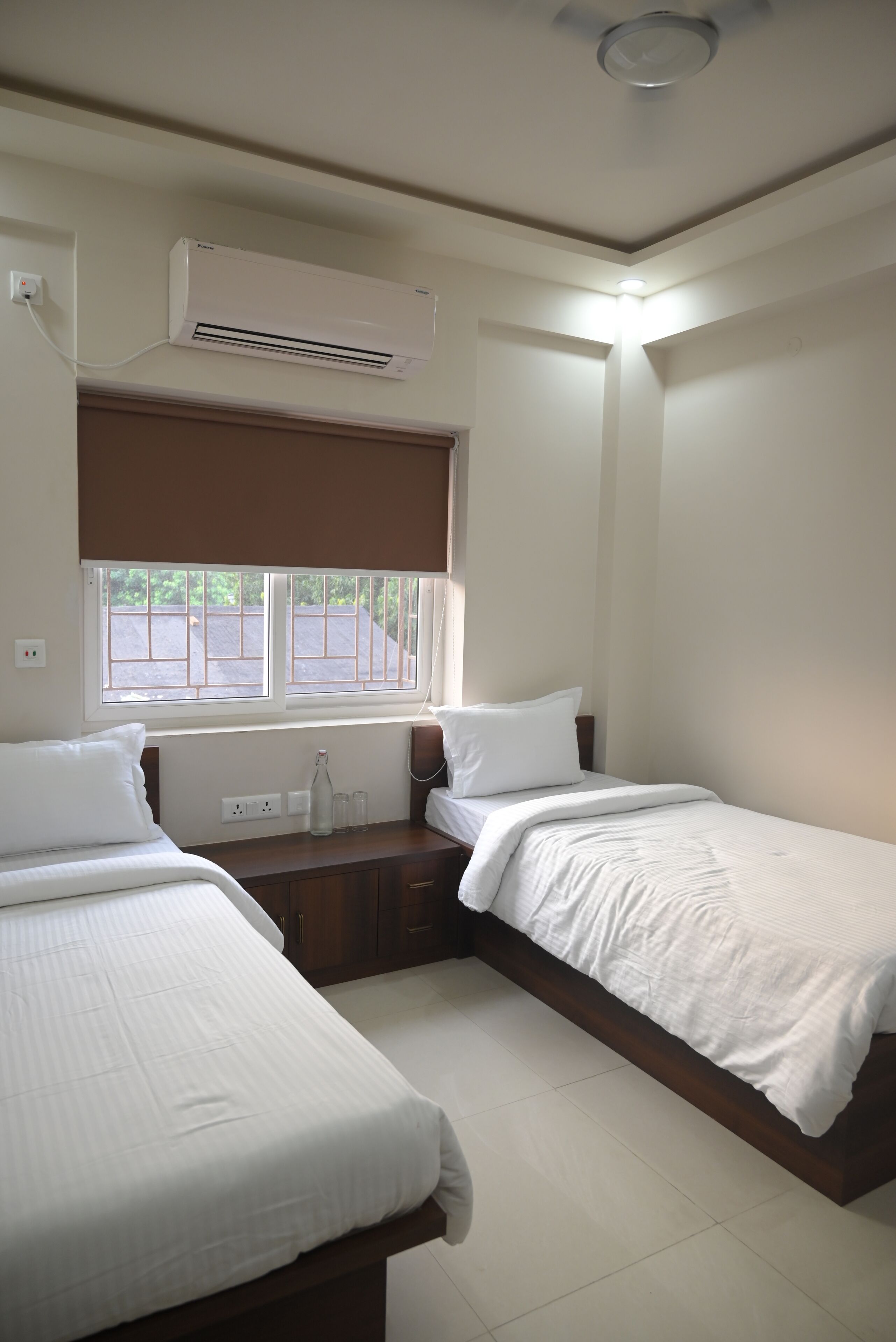 Hotelkamer met twee bedden, een raam met zonwering, een airconditioning en een nachtkastje met een fles en glazen.