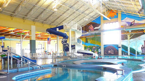 Ein beheiztes Hallenbecken mit Fliesenrand, einer Wasserrutsche und einem Wandgemälde an der Decke.