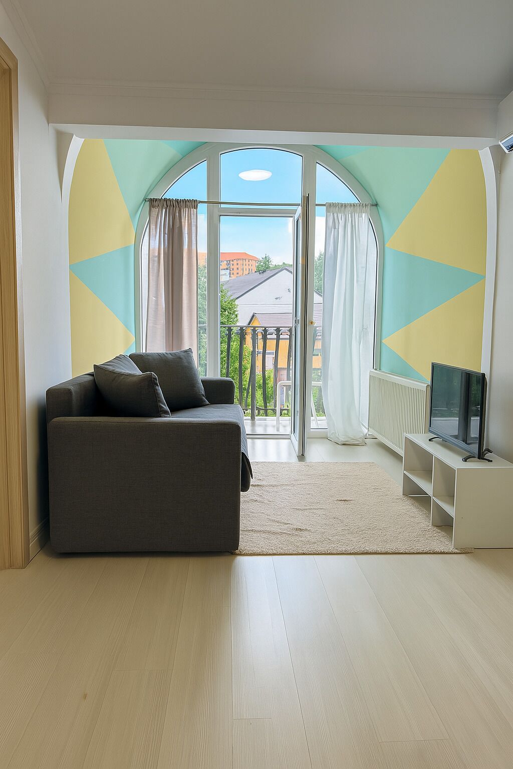 A modern living room with a grey sofa, a flat-screen TV, and a view through large windows to the outside.