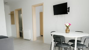 A modern living room with a white dining table, four chairs, a flat-screen TV mounted on the wall, and a vase with flowers on the table.