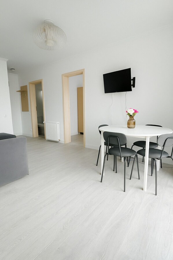 A modern living room with a white dining table, four chairs, a flat-screen TV mounted on the wall, and a vase with flowers on the table. - DD Apartments Iuliu Moldovan (Cluj-Napoca)