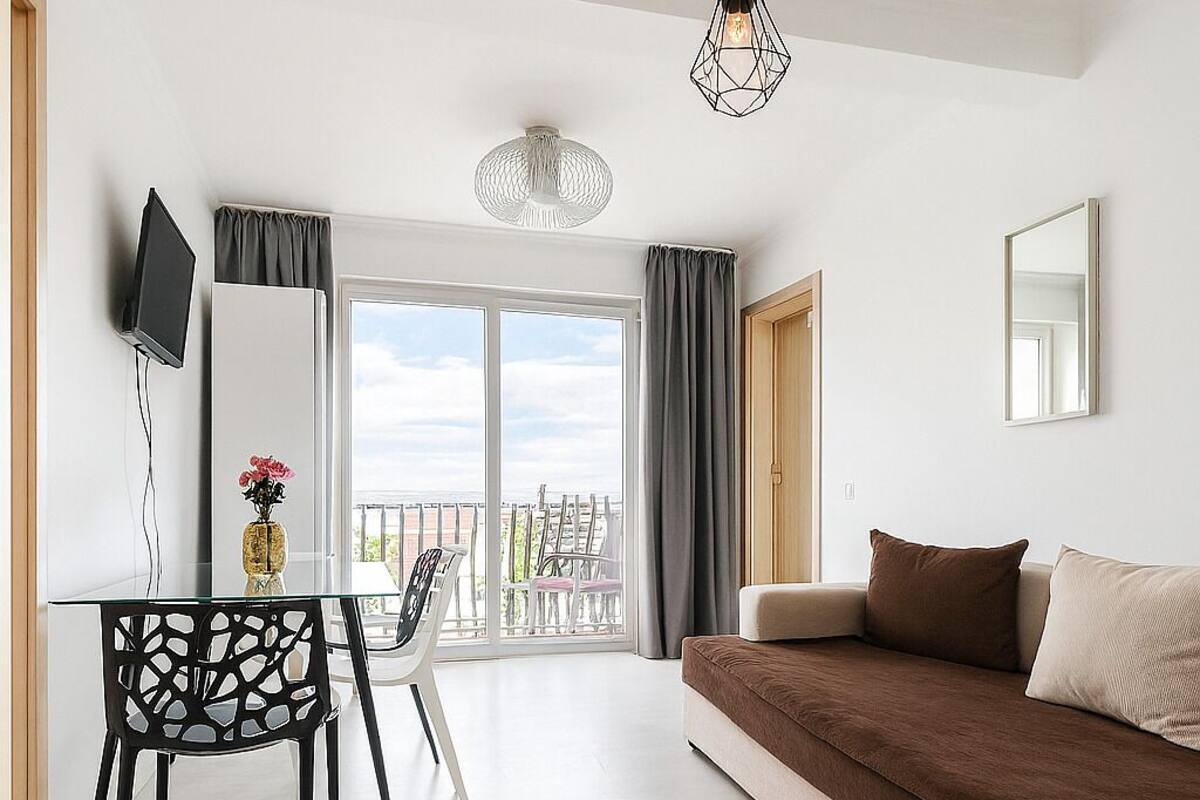 A modern living room with a sofa, a dining table with chairs, a TV mounted on the wall, and a balcony with a view.