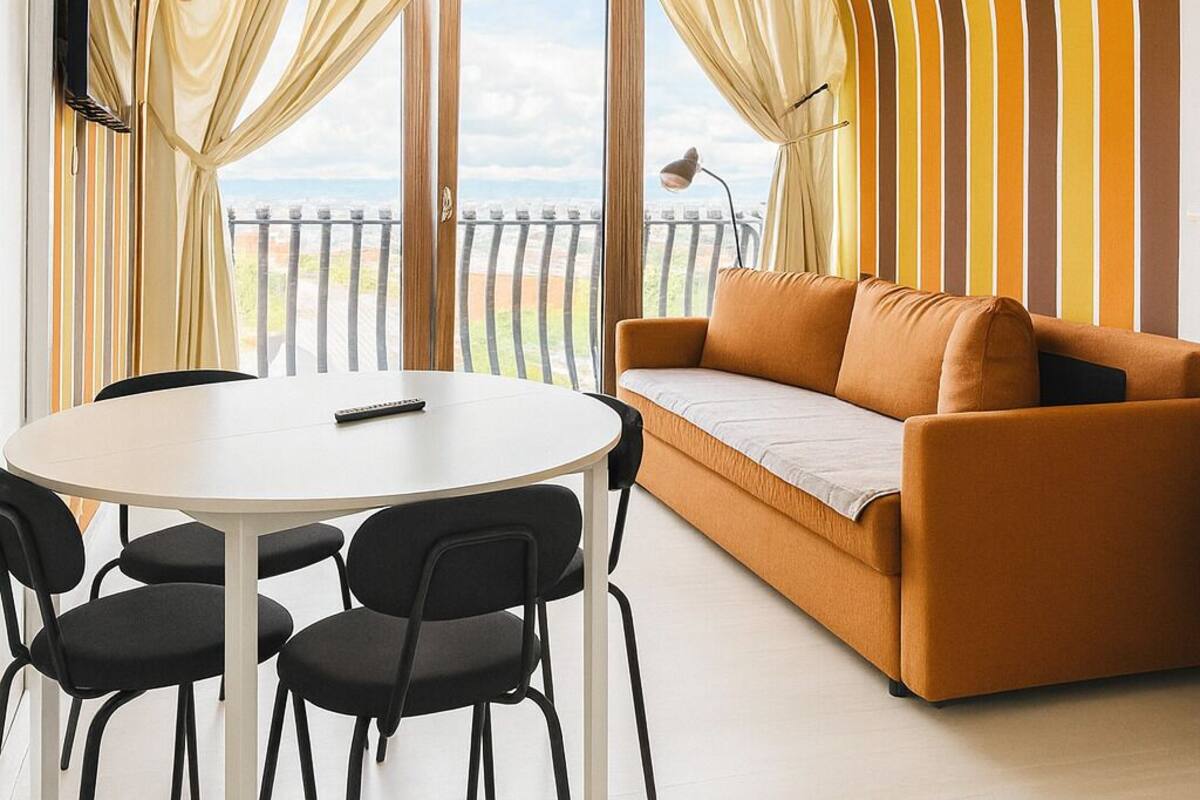 A modern living room with a round table, black chairs, a sofa, and a view through large windows.