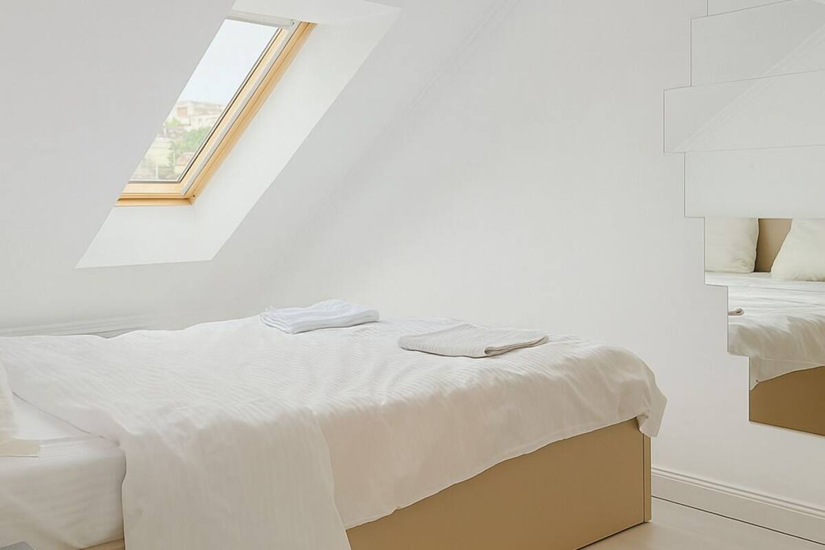 A bedroom with a sloped ceiling, a skylight, a bed with white bedding, a bedside table, and a mirror.