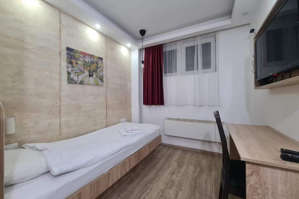 Een kleine, goed verlichte kamer met een enkel bed, een bureau, een stoel, een televisie en een raam met gordijnen.