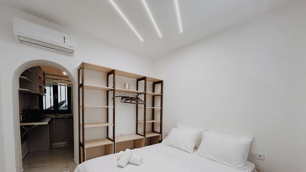 Une chambre à coucher avec un lit, des oreillers blancs, un lit bien fait, un petit tabouret, une armoire avec des étagères et un appareil de climatisation.