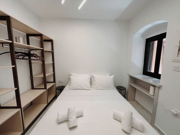 Un dormitorio con cama, dos almohadas, una ventana y estanterías empotradas con ropa colgada.
