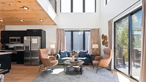 A modern living room with a sofa, coffee table, and chairs, a kitchen with appliances, and large windows with curtains. - Sevierville Retreat • Indoor Pool + Hot Tub + Firepit + Gym | Ridgeview Retreat (Sevierville)