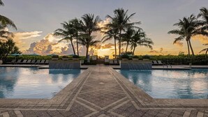 Un área de piscina con un paseo empedrado, sillas de descanso, palmeras y vista al atardecer.