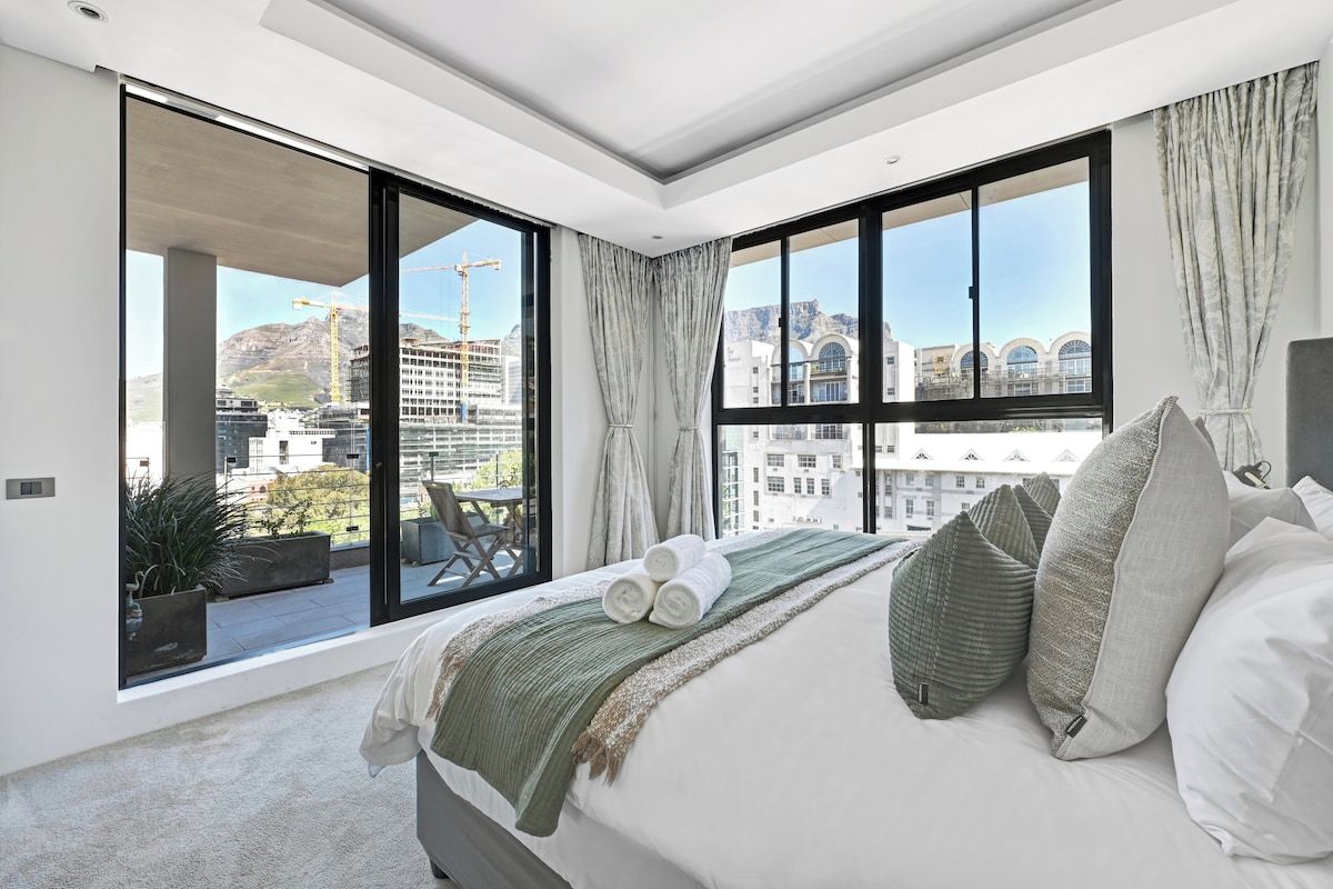 Camera da letto moderna con un grande letto, vista su un cantiere e un balcone con posti a sedere all'aperto.
