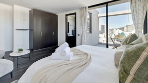 Una habitación de hotel moderna con una cama grande, una pequeña mesa redonda, un sofá y vistas a la ciudad a través de amplias ventanas.