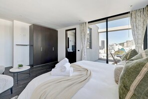 A hotel room with a large bed, a small table, a sofa, and a view of the city through large windows.