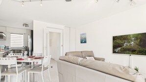 A modern living room with a beige sofa, a dining table with chairs, and a kitchen in the background. - House in residence, pool, 2 adults/2 children (Le Croisic)