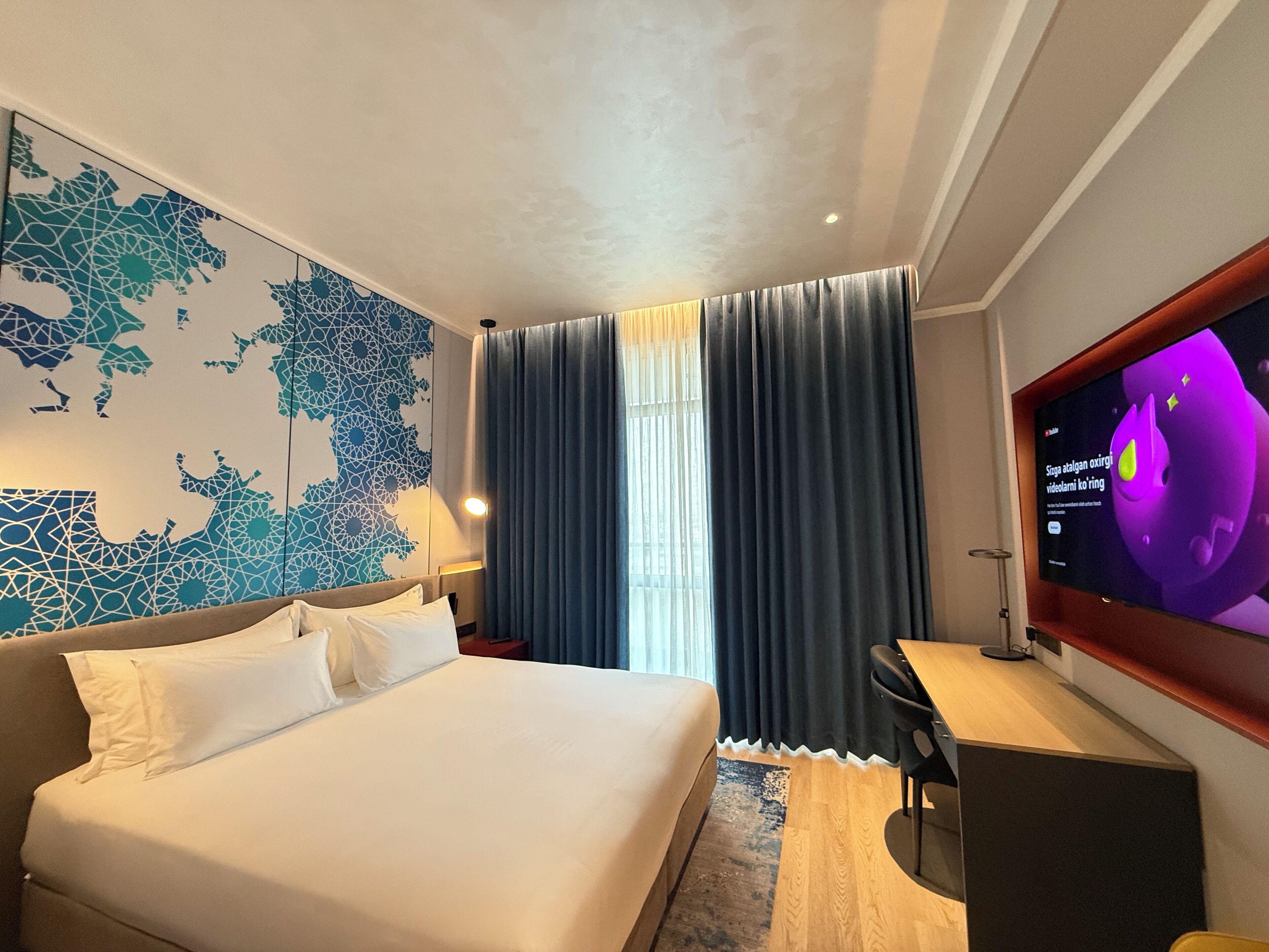 A modern hotel room with a large bed, a desk, a TV, and a world map mural on the wall.