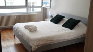 A neatly made bed with white linens and two dark pillows, a bedside lamp, and a view of the outside through large windows.