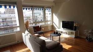 A modern living room with a sofa, armchair, and TV stand, large windows with a view of buildings, and a radiator.