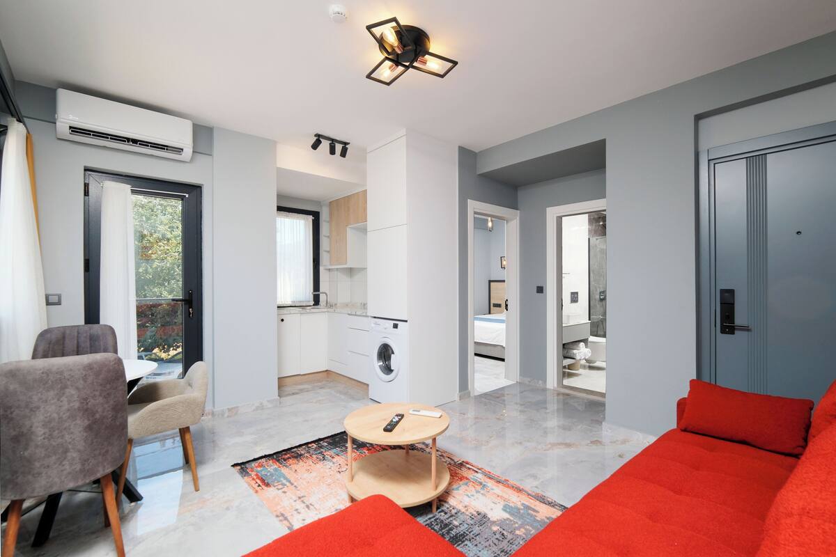 A modern living room with a sofa, a coffee table, and a dining area. There is a kitchen in the background with a washing machine and a dining table.