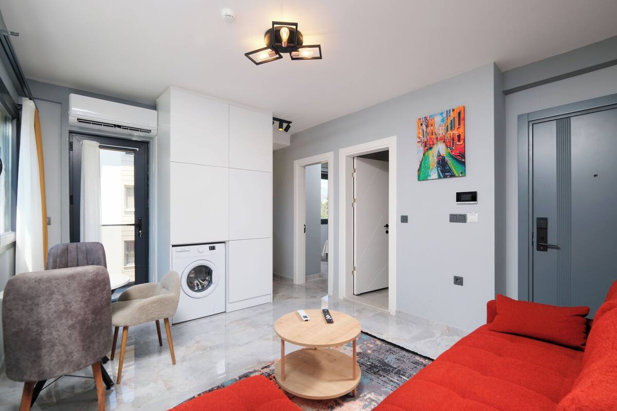 A modern living room with a red sofa, a round wooden coffee table, and a washing machine in the background.