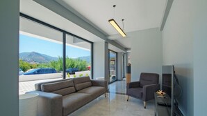 Una sala de estar moderna con un sofá, sillones y un televisor. Amplias ventanas ofrecen vistas a un estacionamiento y montañas.