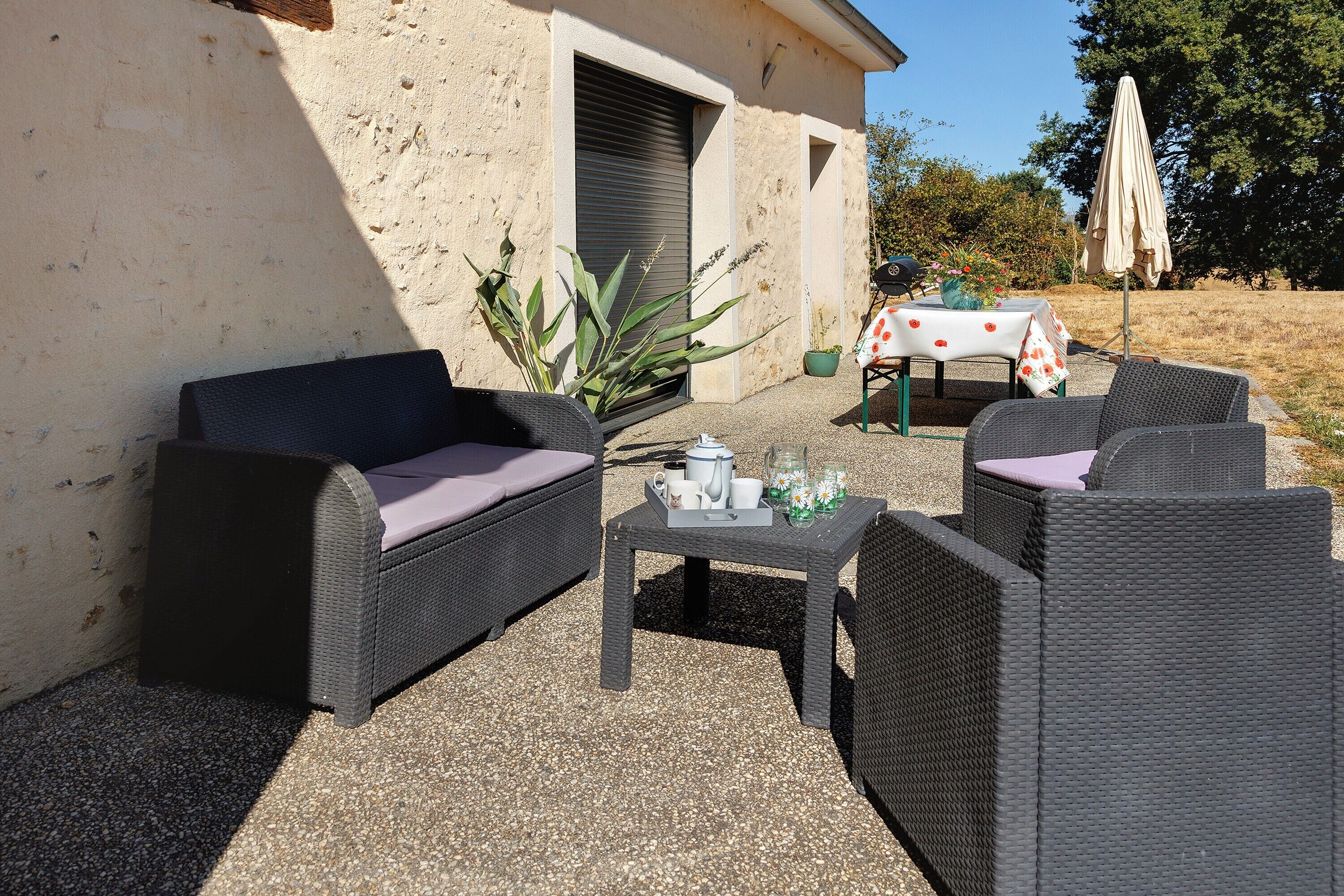 Un espace extérieur aménagé avec des meubles en osier, une table ornée d’un vase de fleurs et un bâtiment dont les volets sont fermés.