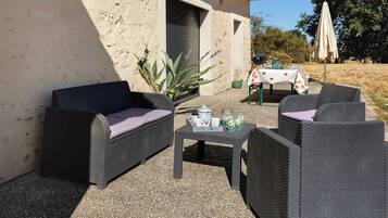 Un espacio al aire libre con mobiliario de mimbre, una mesa con un jarrón de flores y un edificio con una persiana cerrada.