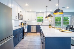 Una cocina moderna con armarios blancos, una isla grande, electrodomésticos de acero inoxidable e iluminación colgante.