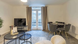Un salon moderne avec une table basse en verre, une table à manger ronde entourée de chaises, un téléviseur à écran plat et une vue dégagée sur l’extérieur grâce à une grande fenêtre agrémentée de rideaux.