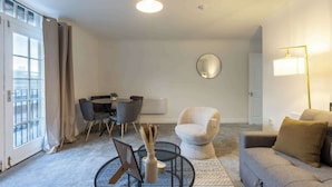 Un salon moderne comprenant un canapé, un fauteuil, une table basse ronde et une table à manger avec des chaises. Un grand miroir orne le mur, et une lampe sur pied est placée dans la pièce.