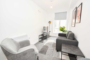 A modern living room with a grey sofa, a grey armchair, a small side table, a floor lamp, and a large window with a view of the outside.