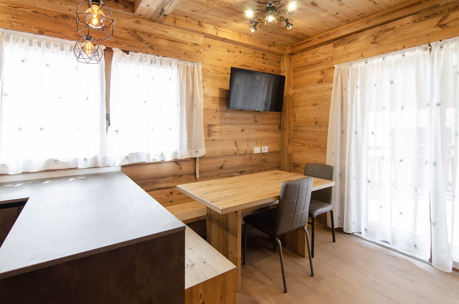 Une cabane en bois avec une table à manger en bois, deux chaises, un téléviseur fixé au mur et un lustre.