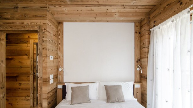 A wooden cabin bedroom with a bed, two pillows, a nightstand, and a window with curtains.
