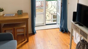 A room with a wooden floor, a desk with a chair, a television, and a large window with blue curtains. - Portuguese Classic Flat at Lisbon Historic Center (Lisboa)