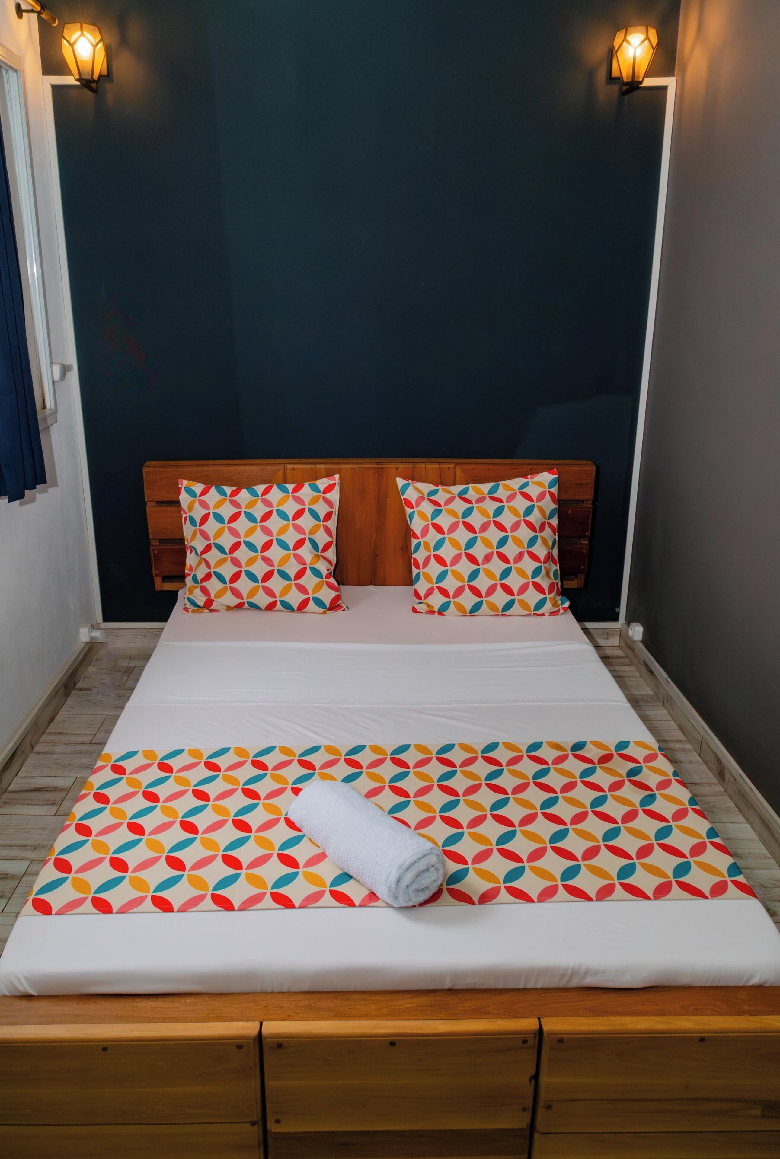 Un letto rifatto con cura, coperta e cuscini coordinati con un motivo geometrico colorato, una testata in legno e due luci fissate al muro.