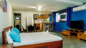 A bedroom with a wooden bed, a desk with a chair, a kitchenette, and a TV. - Cattleya Guest House (Nosy Be)