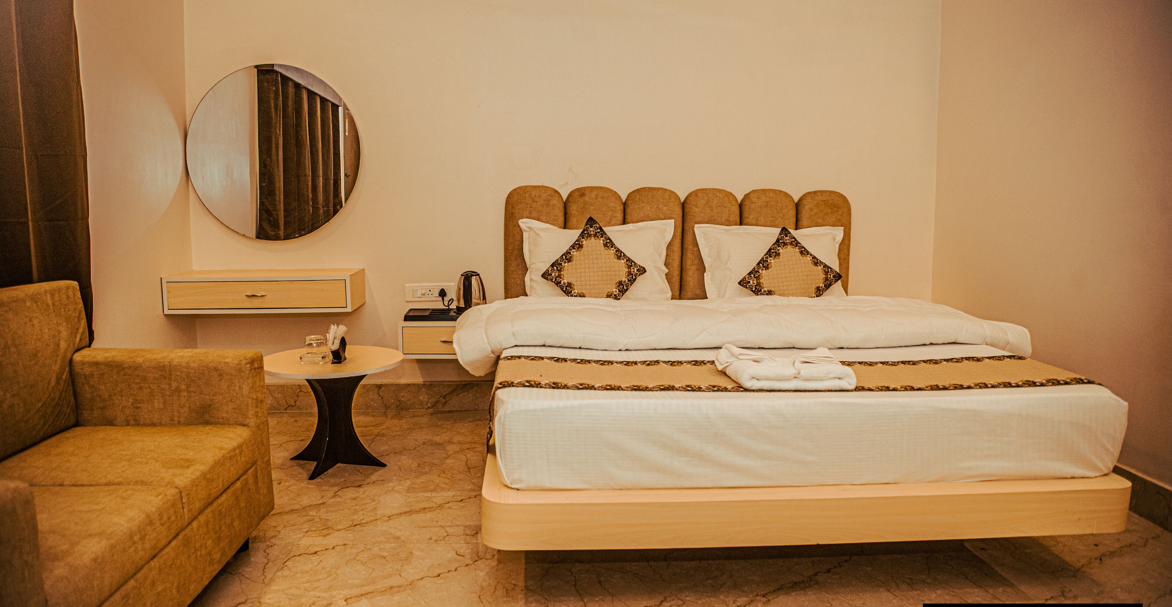 A hotel room with a bed, a round mirror, a bedside table, a small table with a vase, and a sofa.