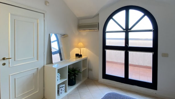 A room with a white door, a white console table with a lamp, a large arched window, and an air conditioning unit on the wall. - Veliero Flexrent - Abyssinia Real Estate (Riccione)