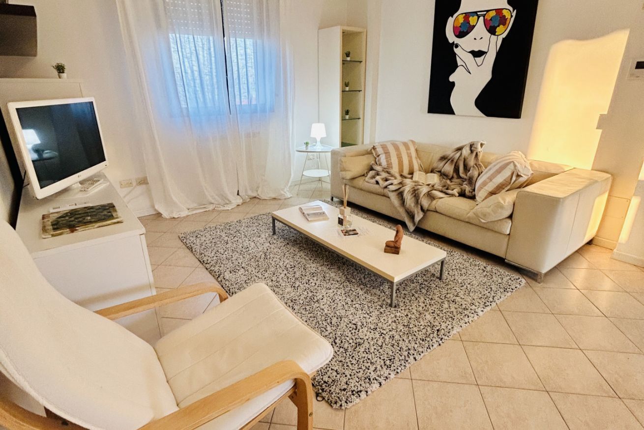 Uma sala de estar moderna com um sofá bege, uma poltrona branca, uma mesa de centro e uma televisão.